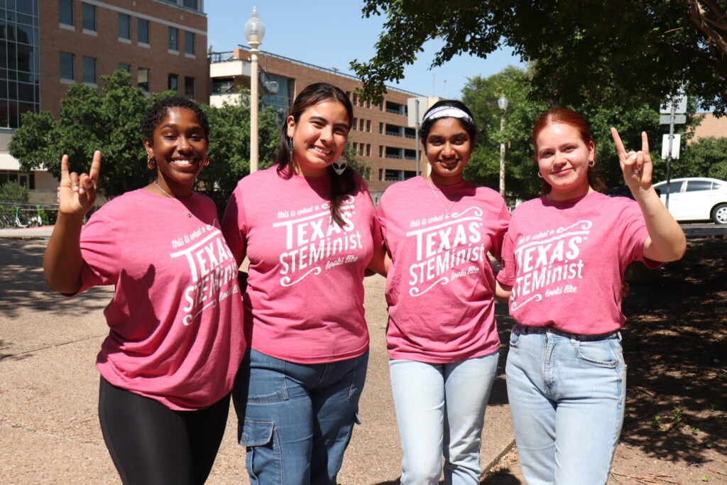 Four undergraduate students wearing pink Texas STEMinist shirts and giving the hook'em horns hand sign.